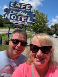 a man and woman pose for a selfie in front of a cafe motel sign